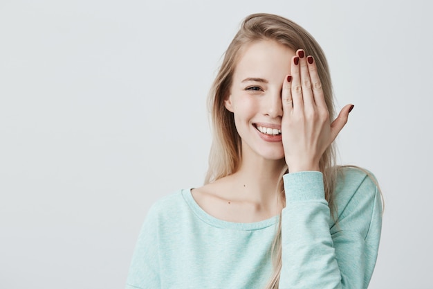 Alegre mujer caucásica con cabello largo teñido, con suéter azul claro, cerrando los ojos con la mano. Feliz mujer positiva con buen humor y juguetón.