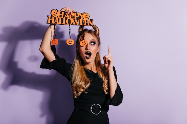 Alegre mujer caucásica en elegante traje negro posando en halloween. Encantadora bruja de pie en la pared púrpura.