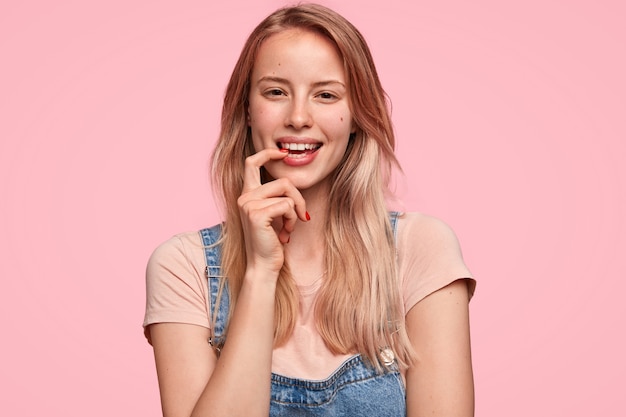 Alegre mujer caucásica muerde el dedo índice, mira con alegría, tiene una expresión feliz, está de buen humor, vestida con ropa informal, se para contra la pared rosa. Personas, emociones, concepto de estilo de vida