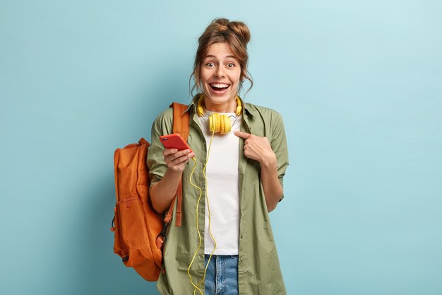 Alegre mujer caucásica se señala a sí misma, pregunta por qué yo, tiene una expresión facial feliz, sostiene el teléfono celular conectado a auriculares estéreo