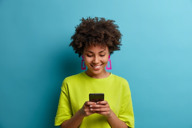 Alegre mujer complacida con cabello rizado sostiene el teléfono móvil y envía mensajes de texto con amigos en las redes sociales, usa una aplicación especial, mira videos interesantes, aislados en la pared azul. Personas y tecnología