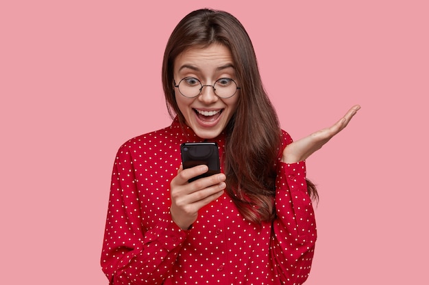 Alegre mujer complacida navega por la red en el teléfono celular, gesticula activamente, hace compras en línea, usa camisa roja