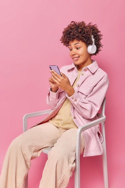 Alegre mujer curiosa centrada en la pantalla del teléfono inteligente escucha la pista de audio en auriculares inalámbricos se sienta en una silla y viste ropa elegante aislada sobre una pared rosa. Personas y tecnologías modernas