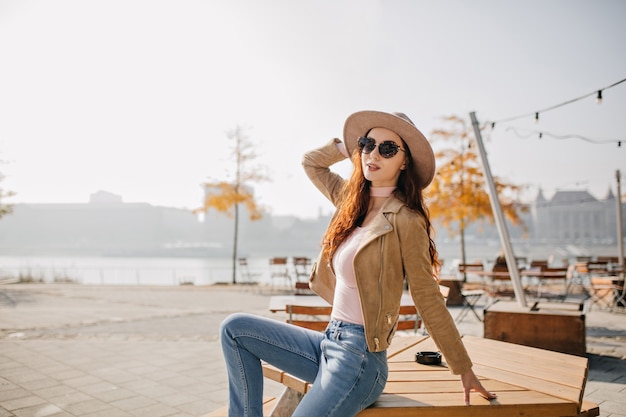 Alegre mujer delgada en jeans sentado en la mesa de madera en el restaurante de la calle