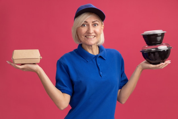 Alegre mujer de entrega rubia de mediana edad en uniforme azul y gorra sosteniendo paquetes de comida de papel y contenedores de comida