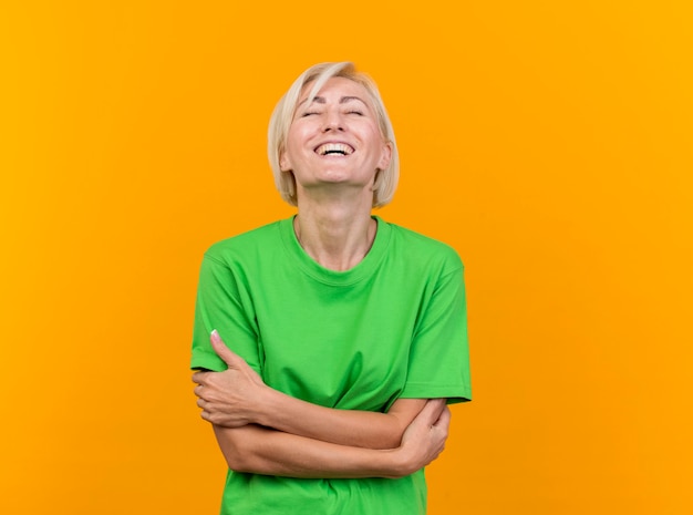 Alegre mujer eslava rubia de mediana edad de pie con postura cerrada y riendo con los ojos cerrados aislado sobre fondo amarillo con espacio de copia