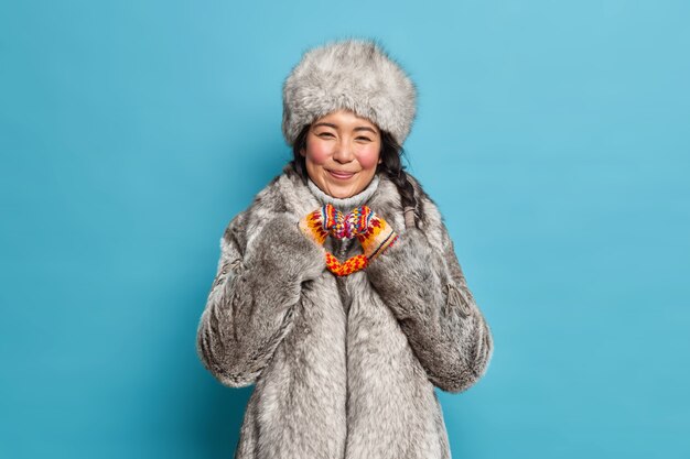 Alegre mujer esquimal forma gesto de corazón expresa amor vestido con ropa de invierno cálido aislado sobre pared azul