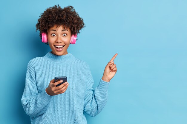 Alegre mujer étnica con cabello afro indica que en la esquina superior derecha sostiene un celular moderno escucha música a través de auriculares estéreo vestida con un suéter tejido