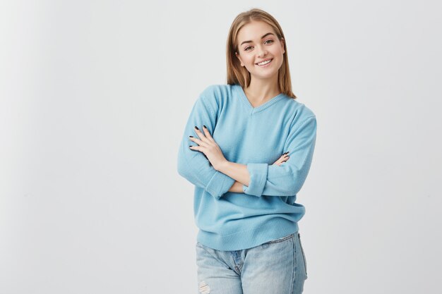 Alegre mujer europea con una apariencia atractiva que sonríe ampliamente con los dientes felices de encontrarse con sus amigos. Estudiante mujer sonriendo a la cámara alegrando su éxito, de pie con los brazos cruzados.