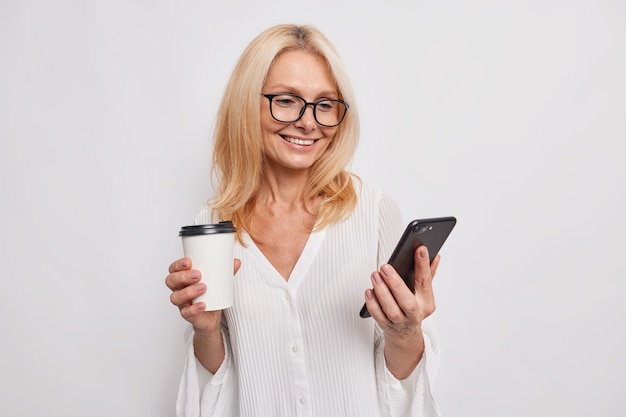 Alegre mujer europea bebe café de una taza para llevar sostiene el teléfono inteligente usa conexión gratuita a Internet durante el descanso sonríe suavemente usa anteojos y elegante blusa aislada sobre una pared blanca