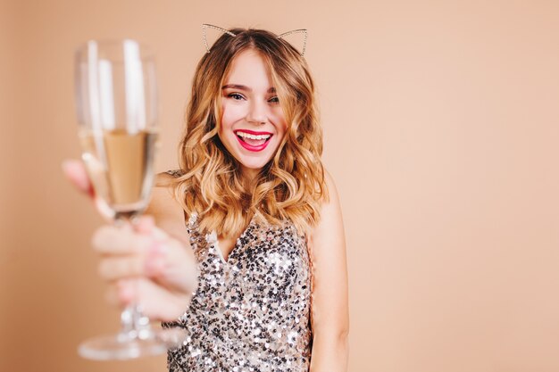 Alegre mujer europea con cabello rizado rubio levantando copa de vino con sonrisa en la pared de luz