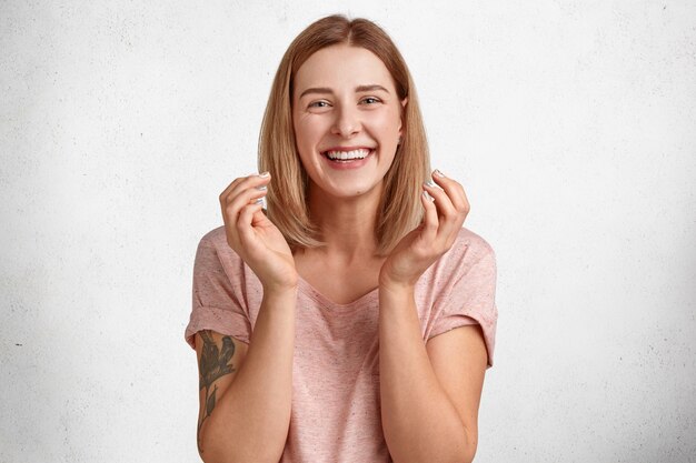 Alegre mujer europea con peinado ondulado, agarra las manos con felicidad, tiene una sonrisa con dientes, muestra una sonrisa blanca y brillante