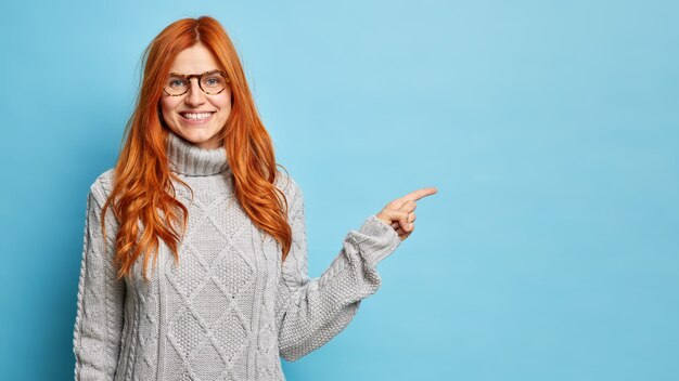 La alegre mujer europea pelirroja con gafas sonríe ampliamente y señala en el espacio de la copia que indica la mejor tienda que usa un suéter de invierno cálido.