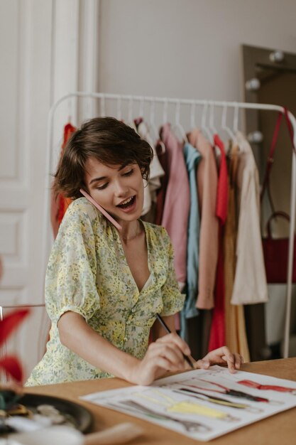 Alegre mujer feliz en vestido floral vneck sostiene pluma y diseña nuevas colecciones de moda Encantadora dama habla por teléfono