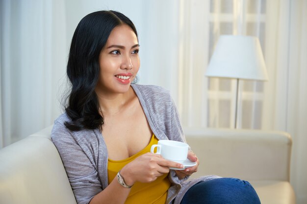 Alegre mujer filipina con bebida caliente en el sofá