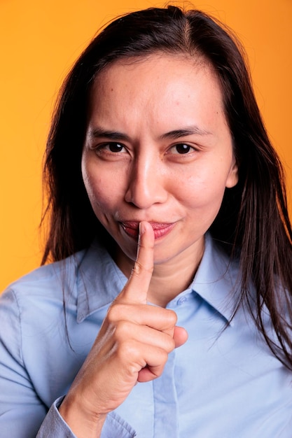 Foto gratuita alegre mujer filipina seria haciendo gesto de silencio poniendo el dedo índice sobre los labios posando en el estudio sobre fondo amarillo. adulto joven haciendo el símbolo shh, expresando confidencialidad