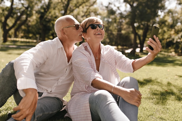Alegre mujer fresca con cabello rubio en blusa moderna a rayas y gafas sentado en la hierba, sonriendo y haciendo selfie con hombre de pelo gris en el parque.
