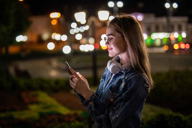 Alegre mujer joven y bonita con anteojos geniales y ropa de moda caminando en la calle de la metrópoli con luces nocturnas disfrutando de canciones de listas de reproducción en línea en auriculares y leyendo sms con buenas noticias en teléfonos inteligentes