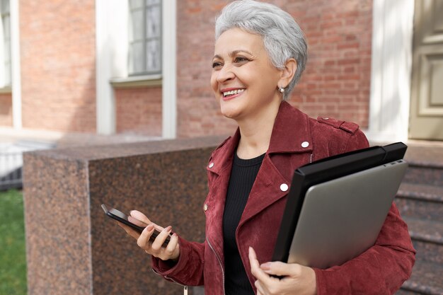 Alegre mujer jubilada posando al aire libre con una computadora portátil y un teléfono inteligente, sonriendo ampliamente, feliz de ver a un amigo