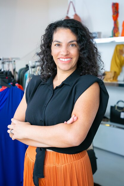 Alegre mujer latina de pelo negro de pie con los brazos cruzados junto a la rejilla con vestidos en tienda de ropa, mirando a cámara y sonriendo. Concepto de asistente de tienda o cliente boutique