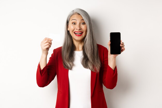 Alegre mujer madura asiática que muestra la pantalla del teléfono inteligente en blanco y tarjeta de crédito, concepto de comercio electrónico.