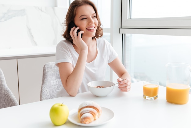 Alegre mujer morena en camiseta blanca hablando por teléfono móvil mientras toma un desayuno saludable en la cocina blanca