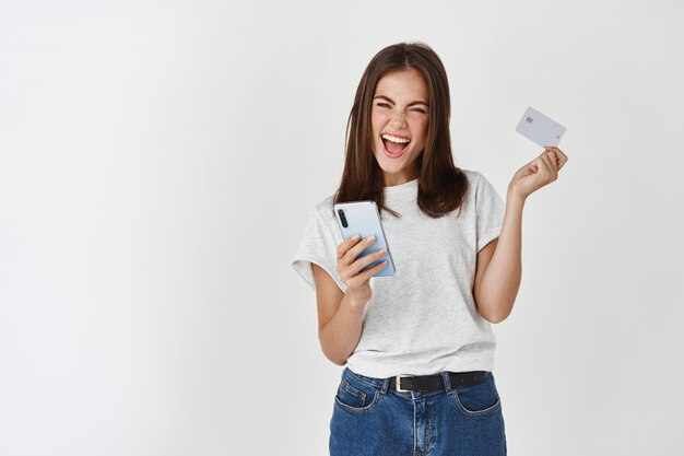 Alegre mujer morena de compras en línea, usando un teléfono inteligente y mostrando una tarjeta de crédito plástica, sonriendo alegre en la pared frontal, blanca.
