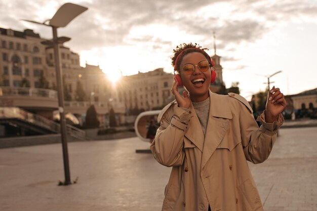 Alegre mujer morena de piel oscura con anteojos y gabardina beige cantando y escuchando música con auriculares al aire libre