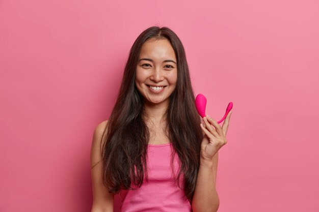 Alegre mujer morena posa con vibrador bluetooth inteligente, usa una aplicación especial en el móvil para mejorar el orgasmo, sostiene la herramienta sexual para mejorar el placer, aislada en la pared rosa. Tecnologías modernas y vida sexual