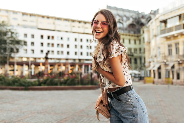 Alegre mujer morena rizada con gafas de sol rosas, pantalones de mezclilla y elegante blusa floral, se ríe y posa de buen humor al aire libre