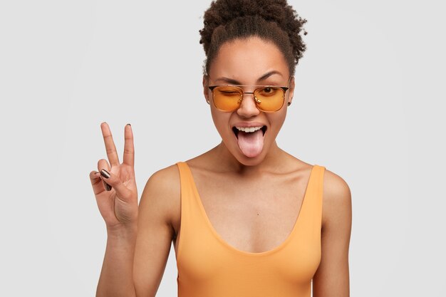 Alegre mujer negra con el signo de la mano de la paz, parpadea y muestra la lengua, tiene el pelo afro rizado peinado en un moño