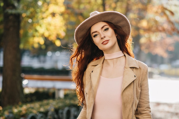Alegre mujer pelirroja de pelo largo con elegante sombrero pasar tiempo libre, explorando la ciudad en día de otoño
