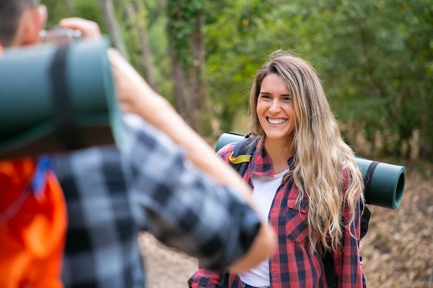 Alegre mujer de pelo largo posando y riendo cuando su novio recortado tomando fotos con la cámara. Excursionistas felices viajando juntos por la naturaleza. Turismo de mochilero, aventura y concepto de vacaciones de verano.