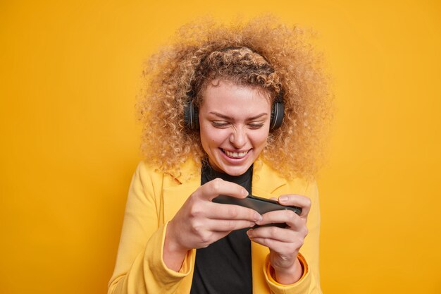 Alegre mujer de pelo rizado adicta a los juegos de video en el teléfono móvil utiliza auriculares inalámbricos concentrados en la pantalla disfruta de los juegos vestida formalmente aislada sobre una pared amarilla vívida