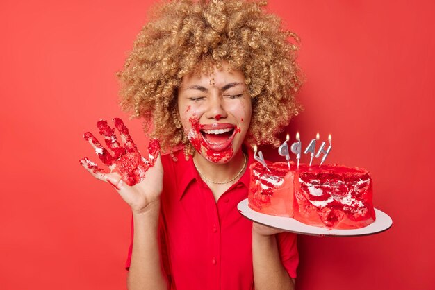 Alegre mujer de pelo rizado se burla de sucia con crema come sabroso pastel dulce con las manos mantiene la palma levantada exclama de alegría viste vestido aislado sobre fondo rojo vivo Comer postre