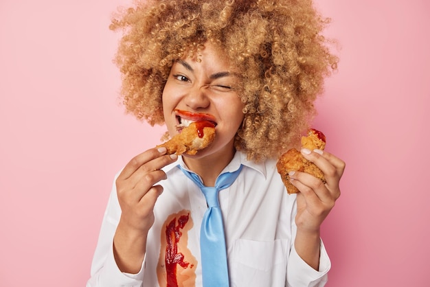 Foto gratuita alegre mujer de pelo rizado come deliciosa comida rápida manchada con ketchup mantiene una nutrición poco saludable tiene el hábito de comer compulsivamente usa camisa blanca y corbata azul aislado sobre fondo rosa