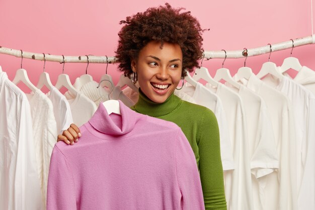 Alegre mujer de pelo rizado compra ropa, sostiene el cuello alto en la percha, se prueba en el vestidor, está ocupada comprando, mira a un lado con una sonrisa, se para en el taller, usa poloneck verde. Estilo de vida de compras