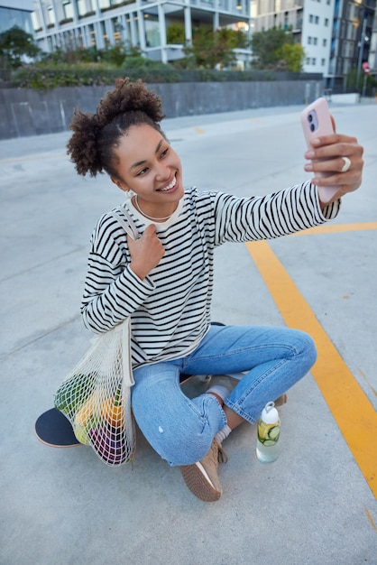 Alegre mujer de pelo rizado usa un jersey de rayas casual y jeans toma selfie en teléfono inteligente se sienta en longboard lleva bolsa de red descansa en la ciudad expresa emociones positivas. Skateboarding de estilo de vida de la gente