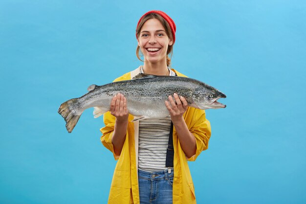 Alegre mujer pescadora en anorak amarillo sosteniendo un enorme pez regocijándose por atraparlo demostrando su trabajo mientras está de pie sobre la pared azul. Gente, afición, recreación y pesca