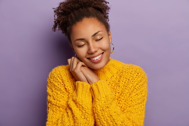 Alegre mujer de piel oscura apoyada en ambas manos, mantiene los ojos cerrados, viste suéter de punto amarillo, modelos sobre la pared violeta del estudio