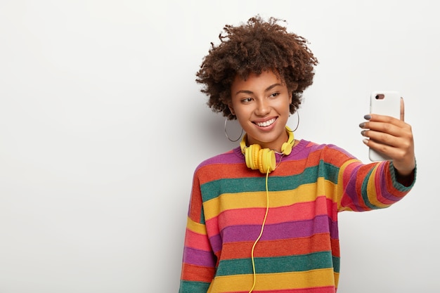 Alegre mujer de piel oscura con cabello oscuro y rizado, hace selfie a través del teléfono inteligente, vestida con un suéter colorido, sonríe suavemente, se para contra el fondo blanco, espacio en blanco a un lado