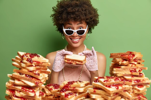 Alegre mujer de piel oscura con cabello rizado sonríe agradablemente come delicioso sándwich