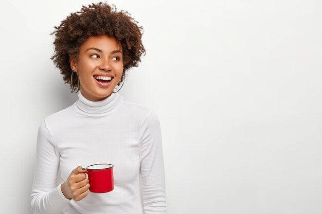 Alegre mujer de piel oscura complacida bebe té de una taza roja, mira en el lado derecho, feliz de tener tiempo libre