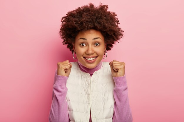 Alegre mujer de piel oscura hace un puñetazo de felicidad, celebra ganar una gran apuesta, usa chaleco blanco, jersey casual, sonríe positivamente, hace gesto de victoria, posa en interiores