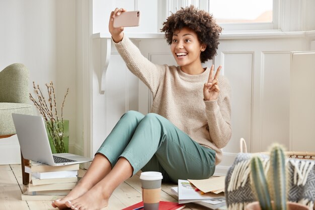 Alegre mujer de piel oscura hace videollamada con teléfono celular, muestra el signo de la paz