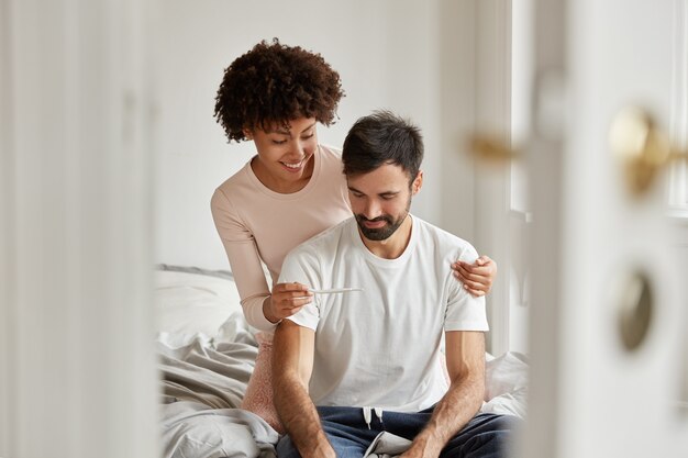 Alegre mujer de piel oscura muestra el resultado de la casa de embarazo en casa a su novio, tiene sonrisas en las caras, se viste con ropa informal, posa en un dormitorio acogedor. La pareja familiar se regocija con la noticia del embarazo juntos.