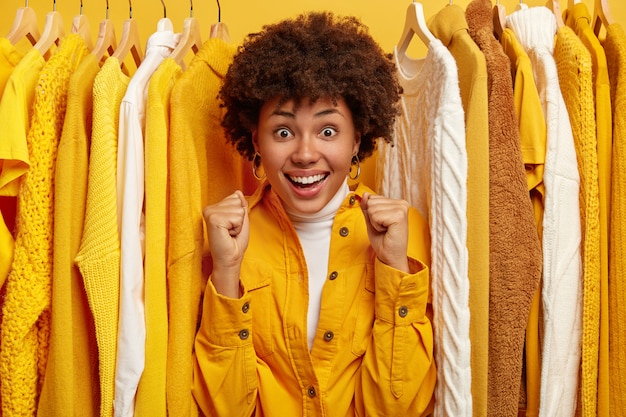 Alegre mujer de piel oscura con peinado afro, aprieta los puños, vestida con ropa de moda, se para cerca de colgar la ropa en rieles