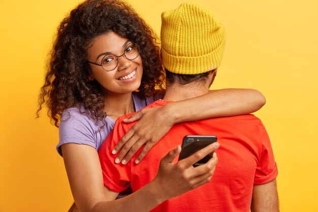 Alegre mujer de piel oscura con peinado afro, usa anteojos redondos, abraza al hombre con sombrero amarillo y camiseta roja, sostiene el teléfono móvil, espera una llamada importante. Personas, tecnología, concepto de relación