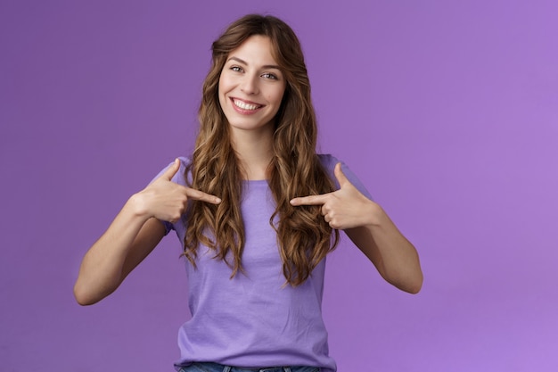 Foto gratuita alegre mujer rizada asertiva profesional motivada apuntando a sí misma en el centro sonriendo ampliamente proponer su propia ayuda quiere participar jactanciosos logros de hablar sobre fondo púrpura.