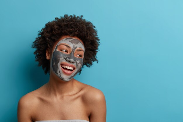 Alegre mujer rizada de piel oscura positiva se para desnuda en el interior, aplica mascarilla de barro de belleza para una piel facial suave y perfecta, se preocupa por la tez, mira con alegría hacia otro lado, aislada en la pared azul.